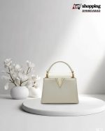 Cream ivory structured handbag with a gold V-shaped accent and top handle, placed on a white round platform with minimal floral decor.